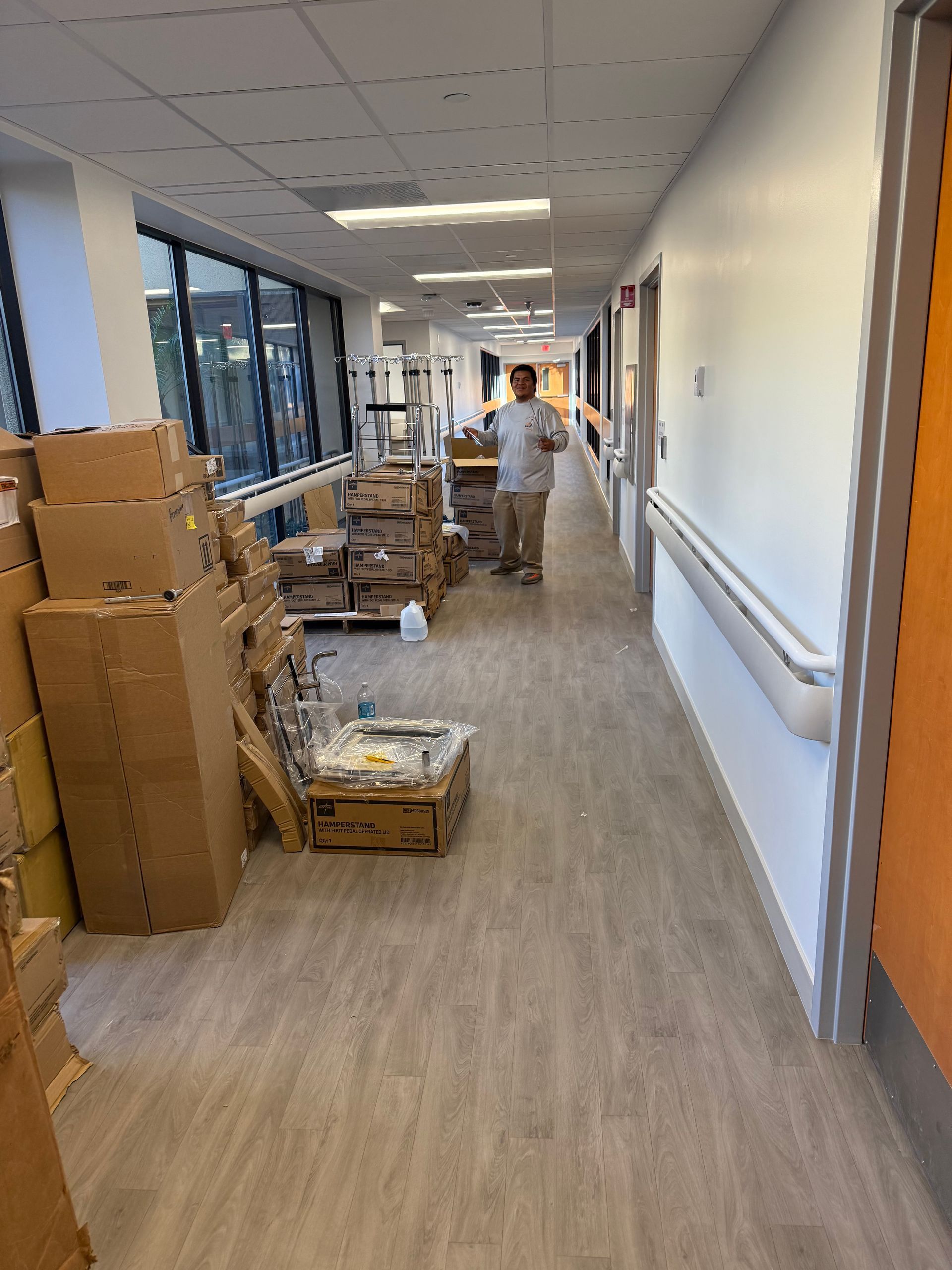 Hallway lined with stacked cardboard boxes and supplies beside large windows and a person walking away