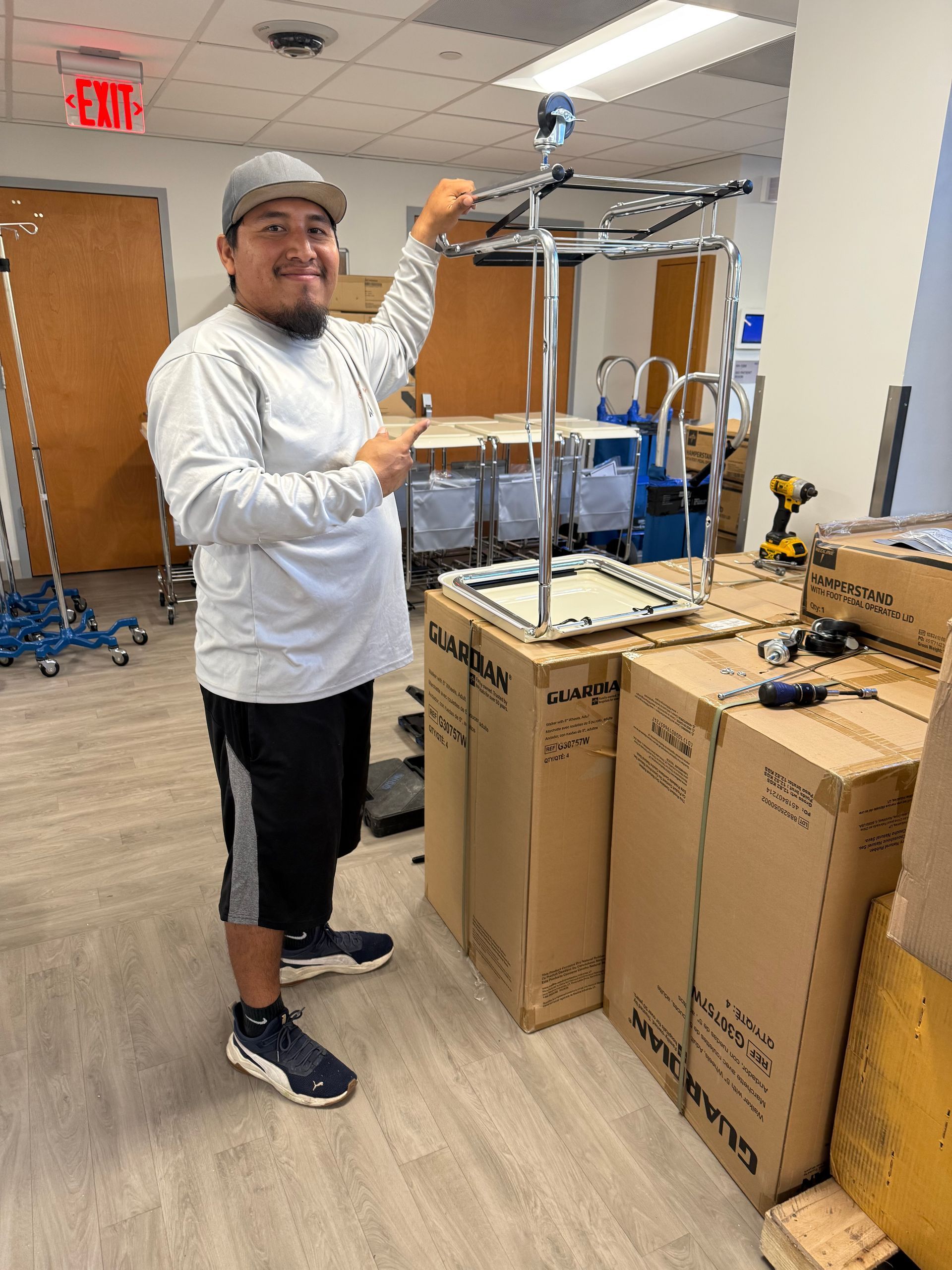 Person in a warehouse stands beside stacked cardboard boxes and a metal frame, holding up a part.