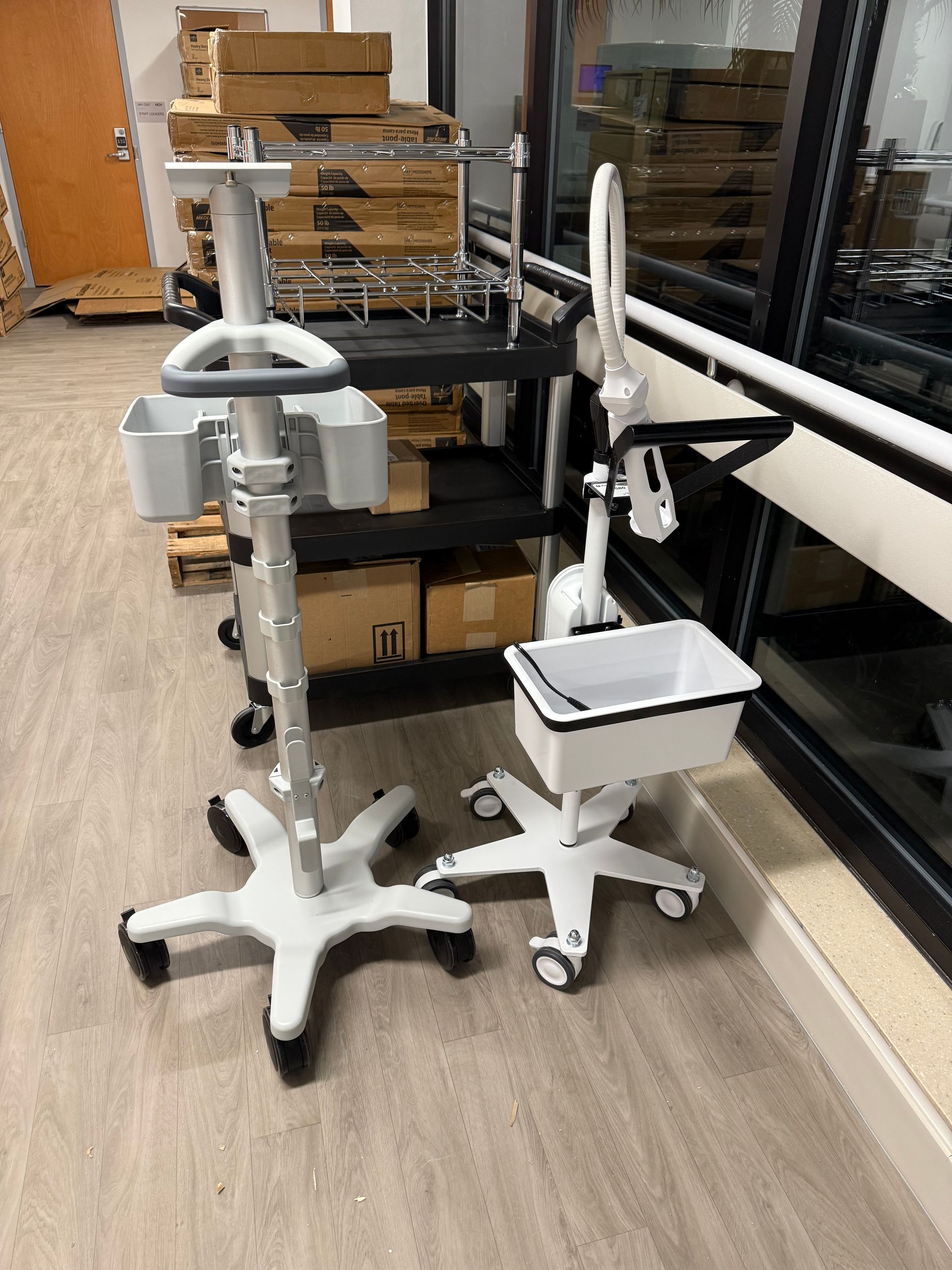 Two white rolling medical stands beside a black cart in a room with boxes and a window.