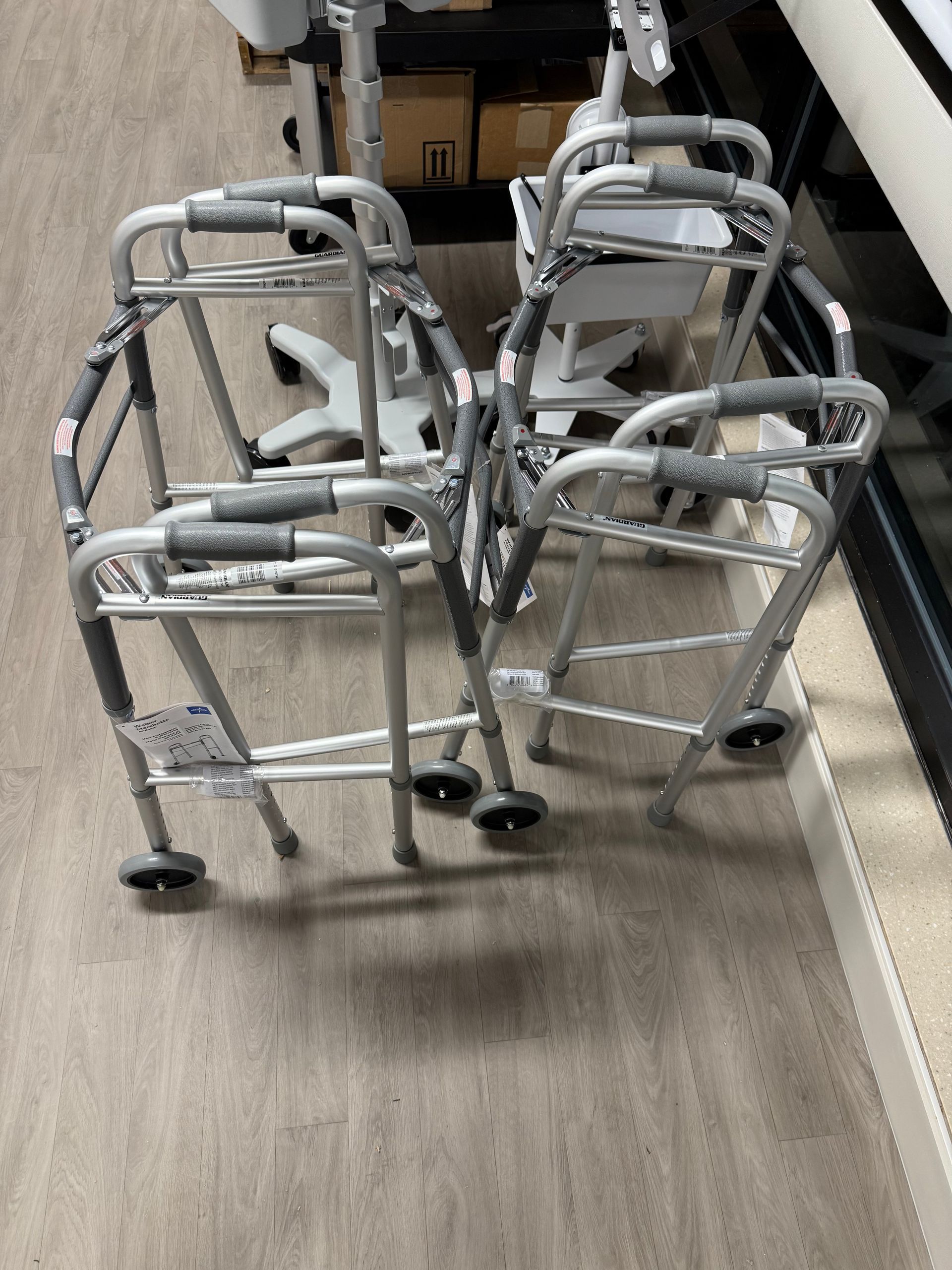 Four silver walkers stacked together on a wood floor in a hallway or storage area