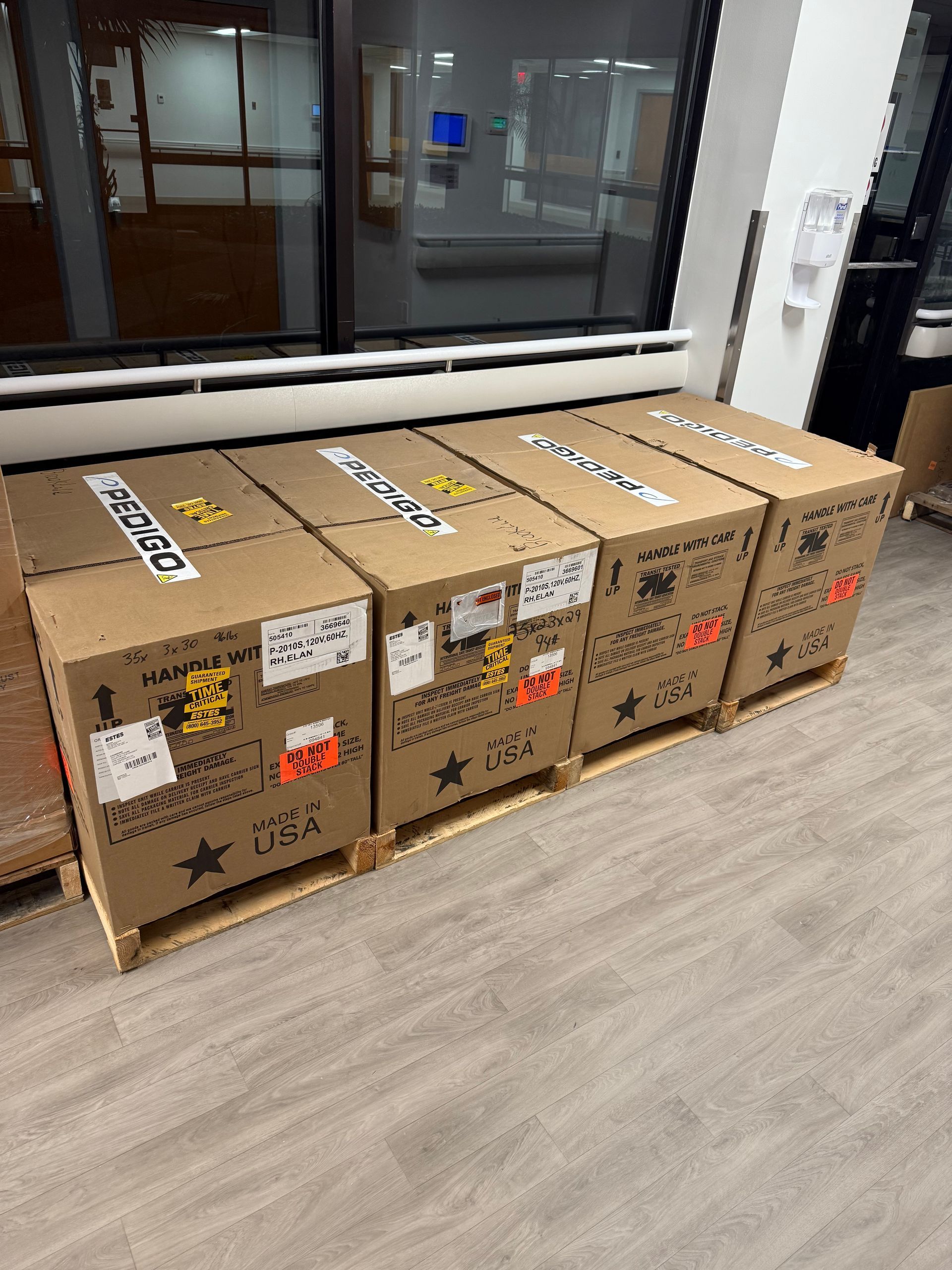 Five cardboard shipping boxes stacked in a row on a floor beside a glass wall.