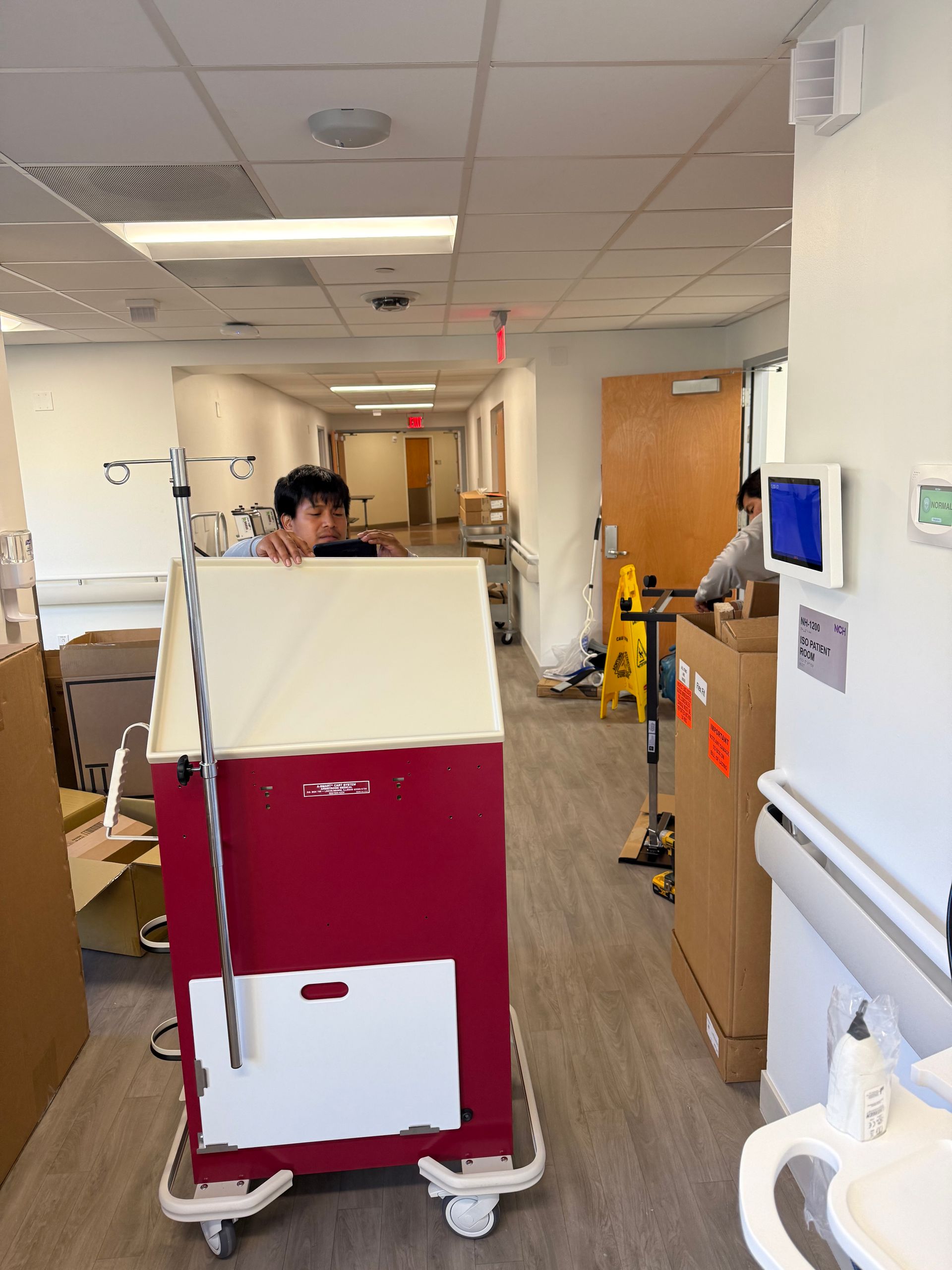 Hospital hallway with a red-and-white cart blocking the path and staff in the background.