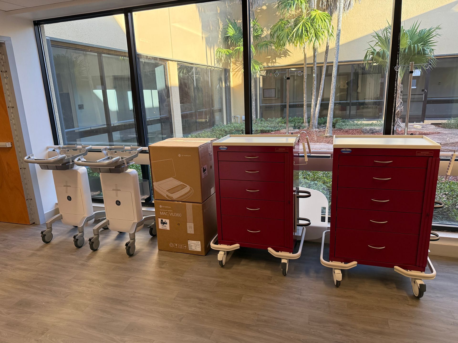 Hospital hallway with two red carts, two stainless-steel carts, and cardboard boxes by glass windows.