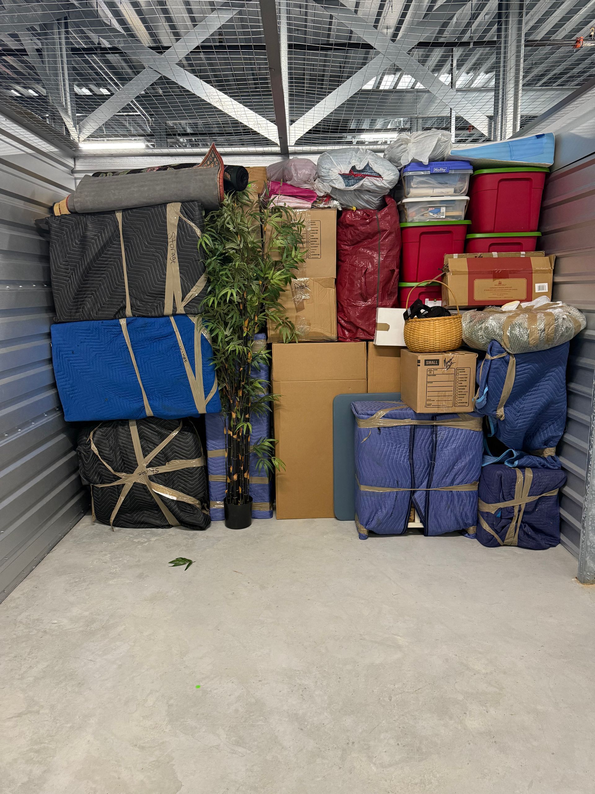 Storage unit packed with wrapped boxes, bags, and a small artificial tree under a metal ceiling