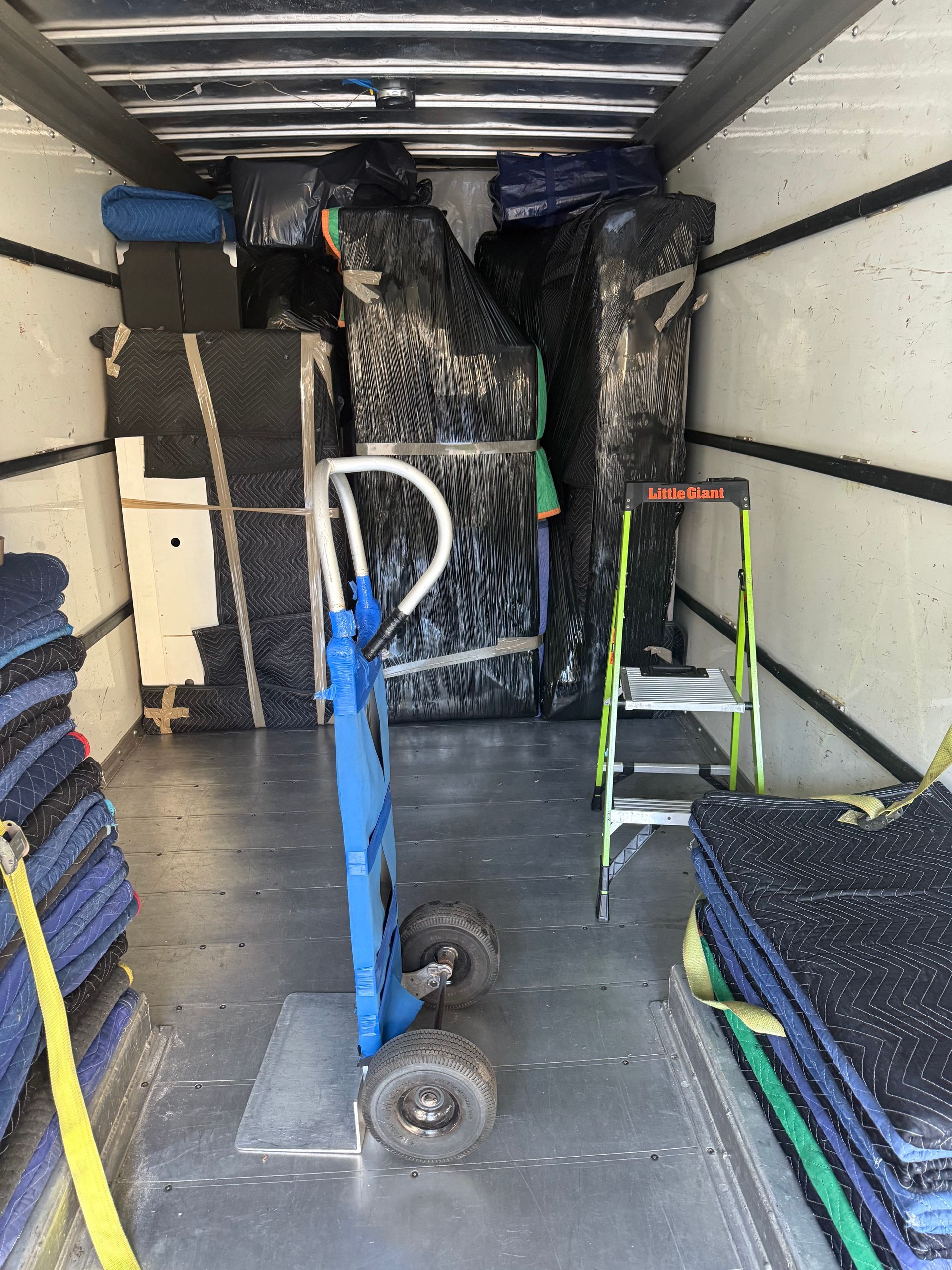 Interior of a cargo trailer with stacked gym mats, a hand truck, and shelving along the walls.