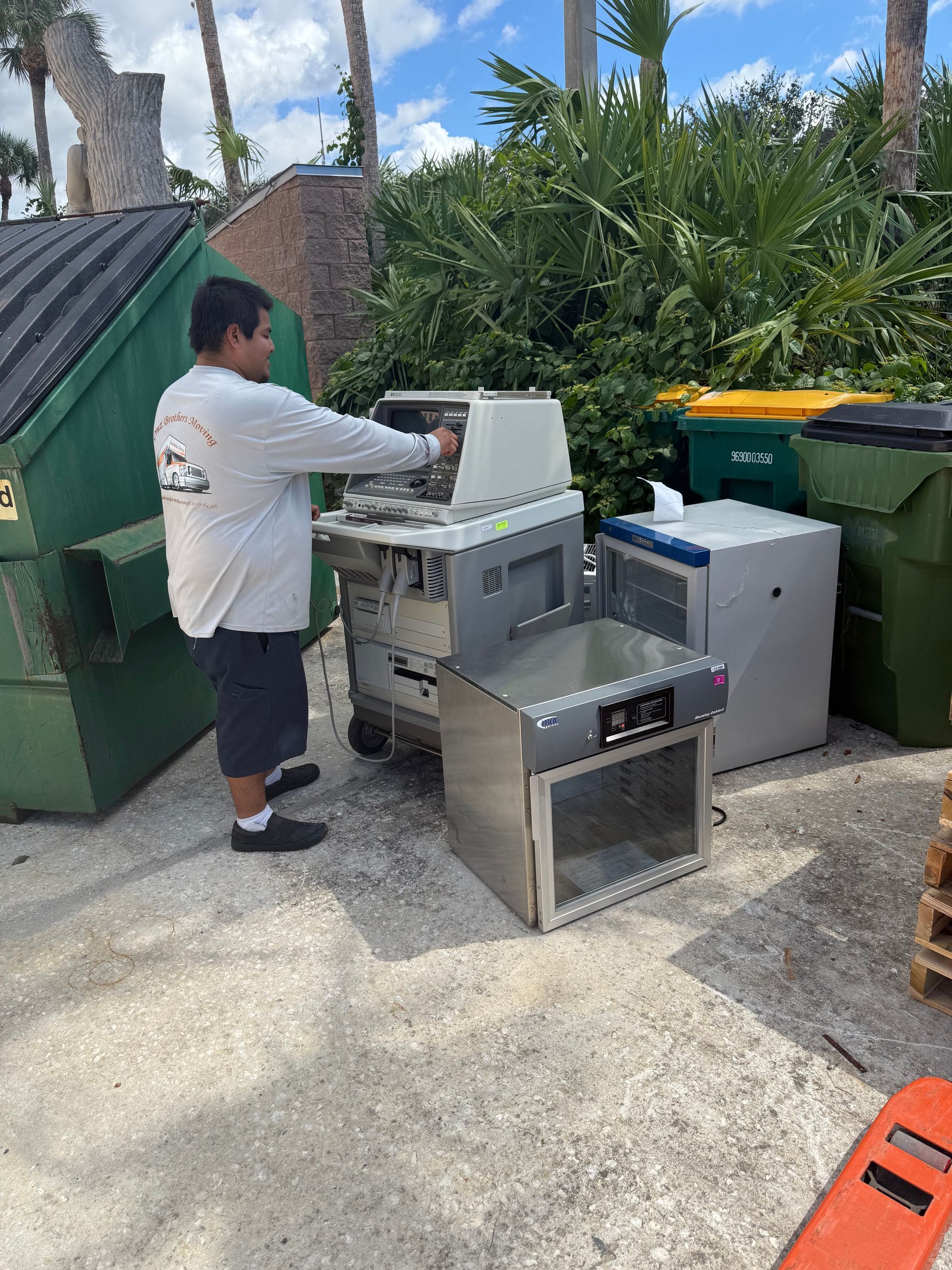 Person loading a stainless steel grill into a cart beside outdoor bins and palm trees