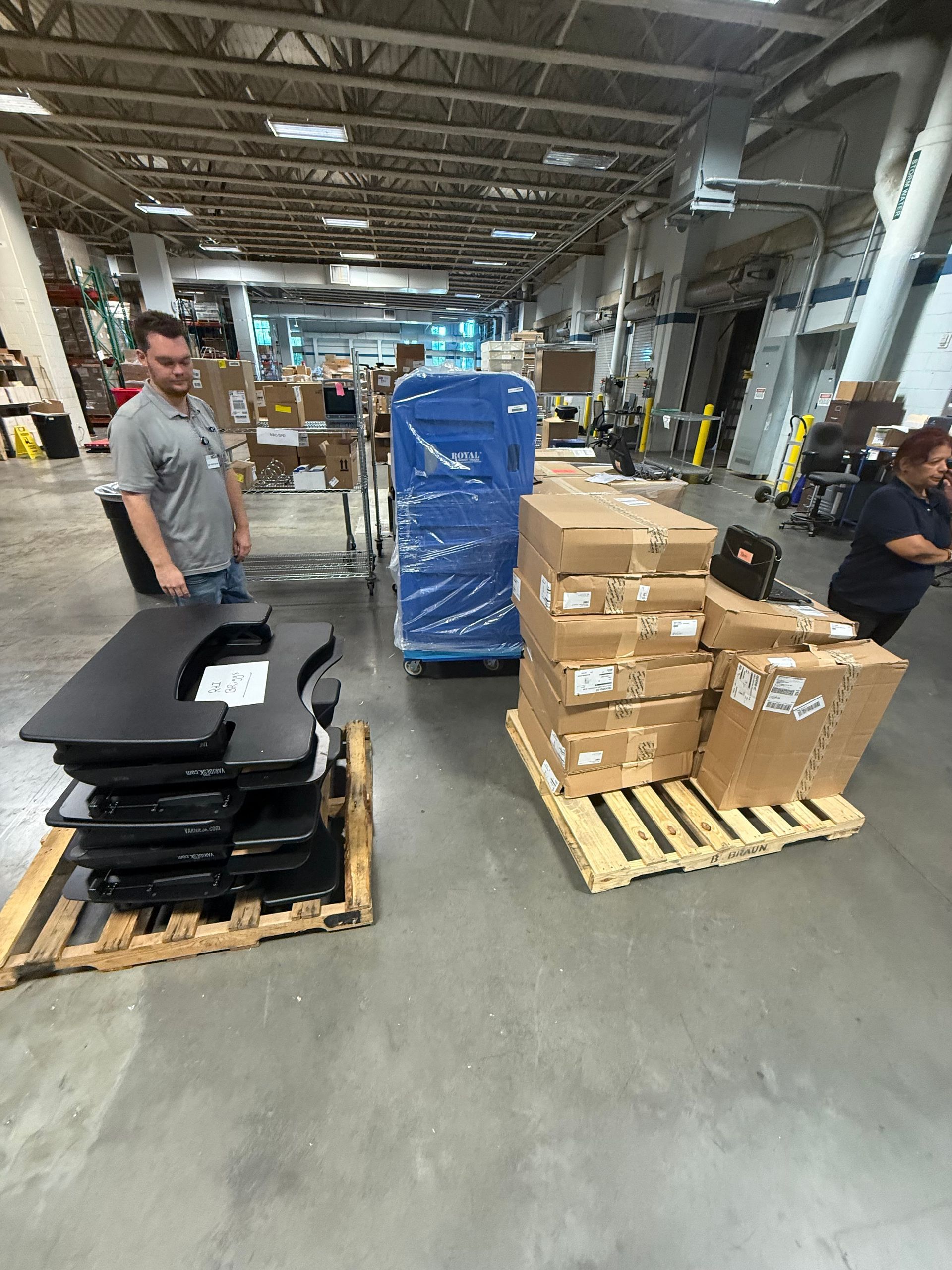 Warehouse aisle with two workers, stacked boxes on pallets, and a blue wrapped pallet in the center.