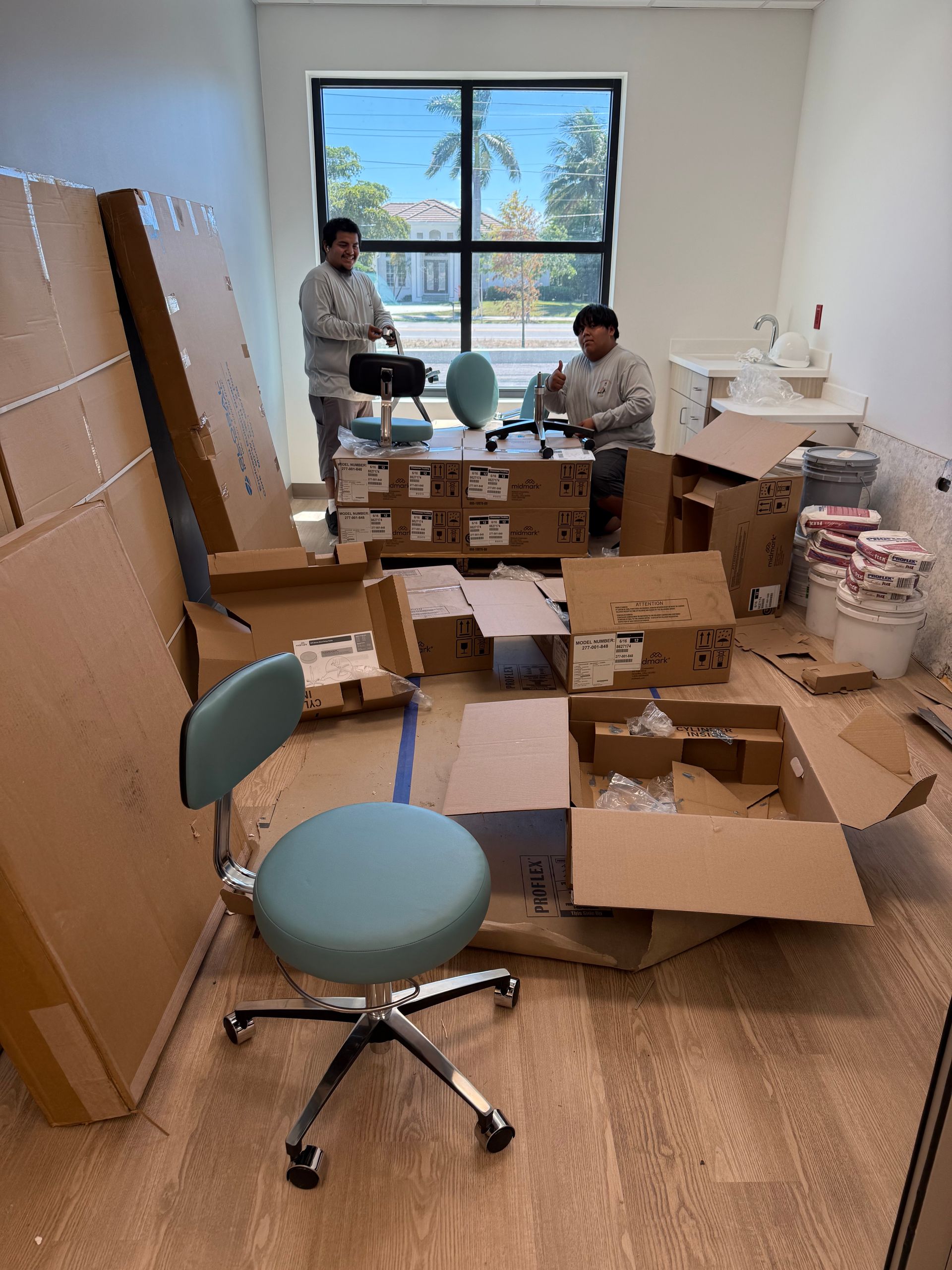 Two people in a cluttered room surrounded by cardboard boxes and packing materials near a window.