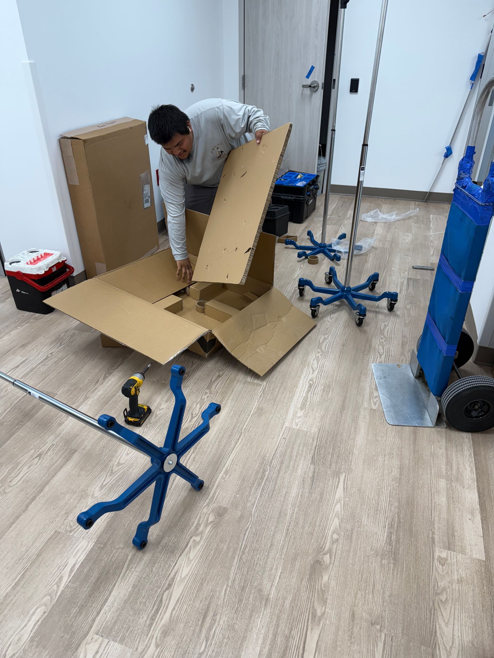 Person assembling furniture among cardboard boxes and blue chair bases in a room with wood flooring.