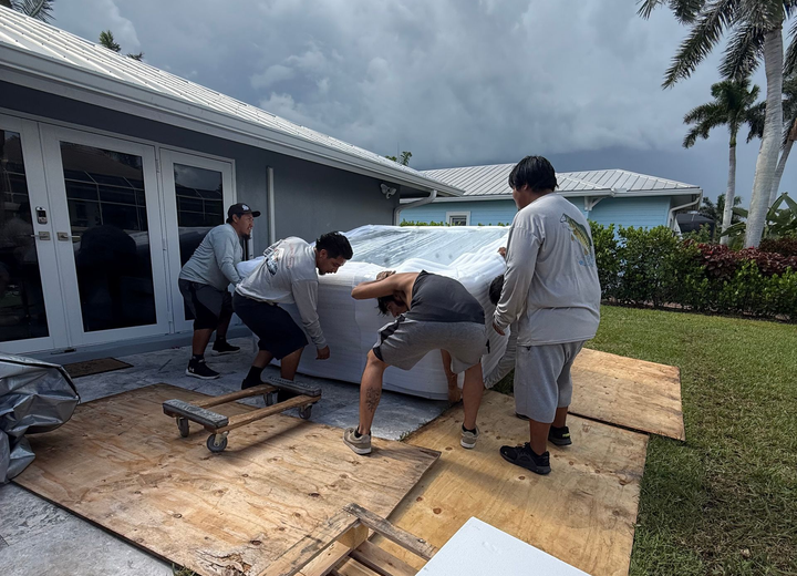 Four workers move a heavy object on plywood outside a white house near the ocean.