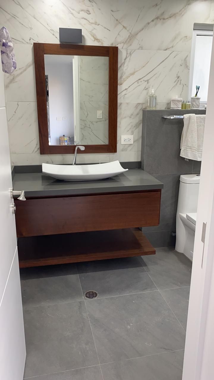 Un baño moderno con tocador de madera, lavabo blanco sobre encimera, espejo rectangular y suelo y paredes revestidos de azulejos grises.
