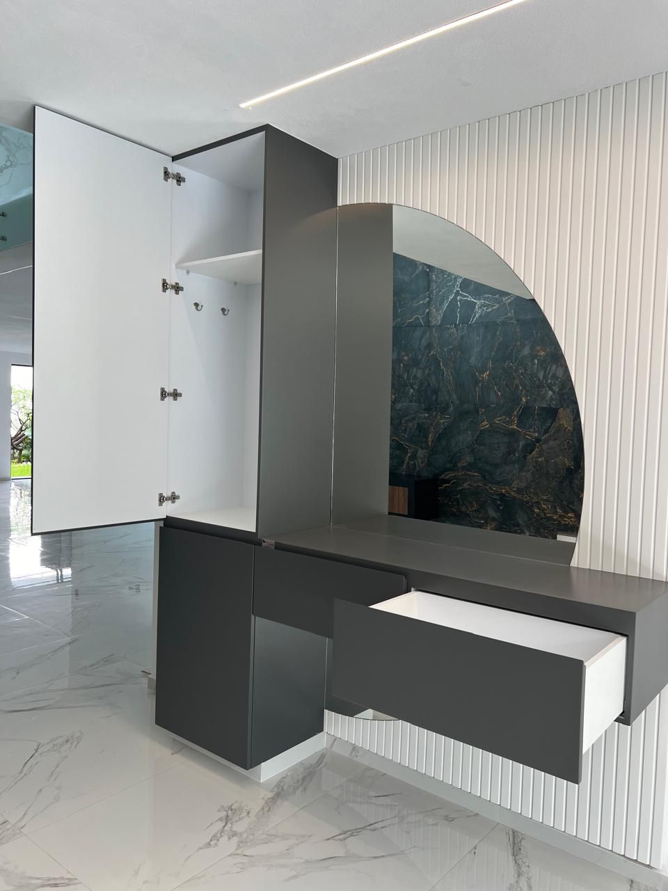 Tocador gris oscuro con cajón abierto y mueble, espejo redondo, pared texturizada blanca.