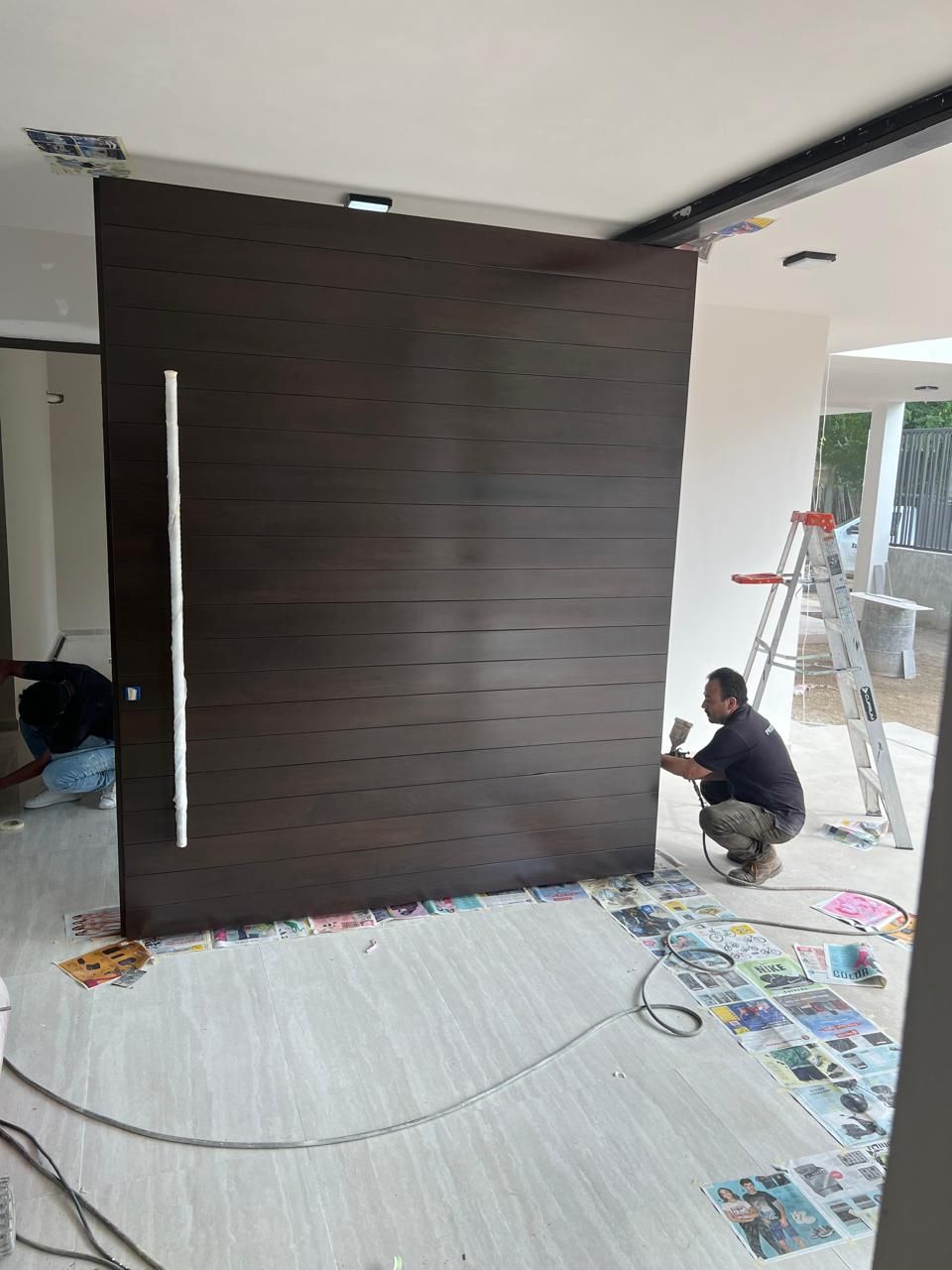 Persona pintando una gran puerta corredera marrón oscuro. Interior con escalera y materiales dispersos en el suelo.