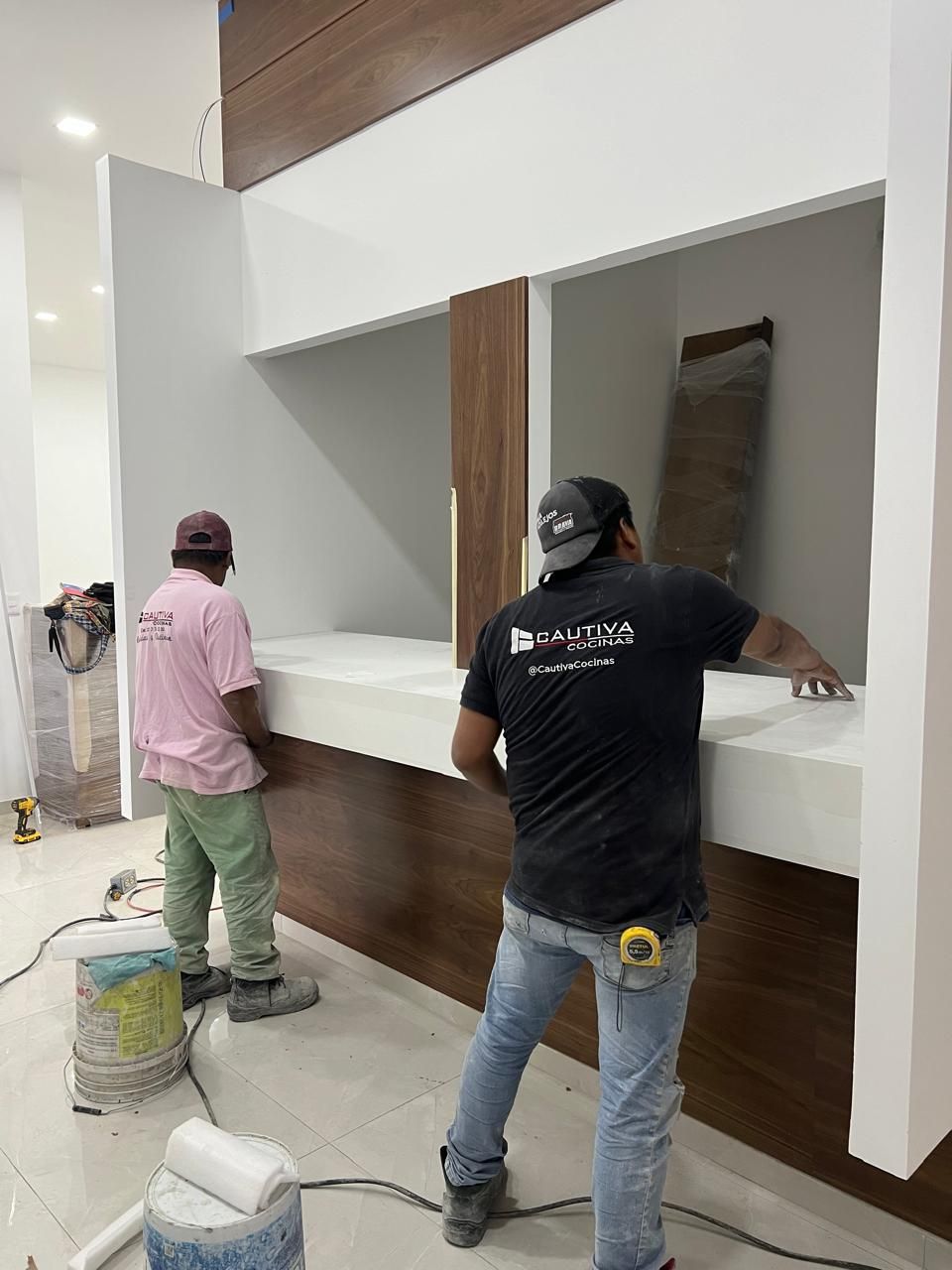 Dos trabajadores instalando un mostrador; uno de rosa, el otro de negro. Revestimiento blanco y de madera en una habitación en construcción.