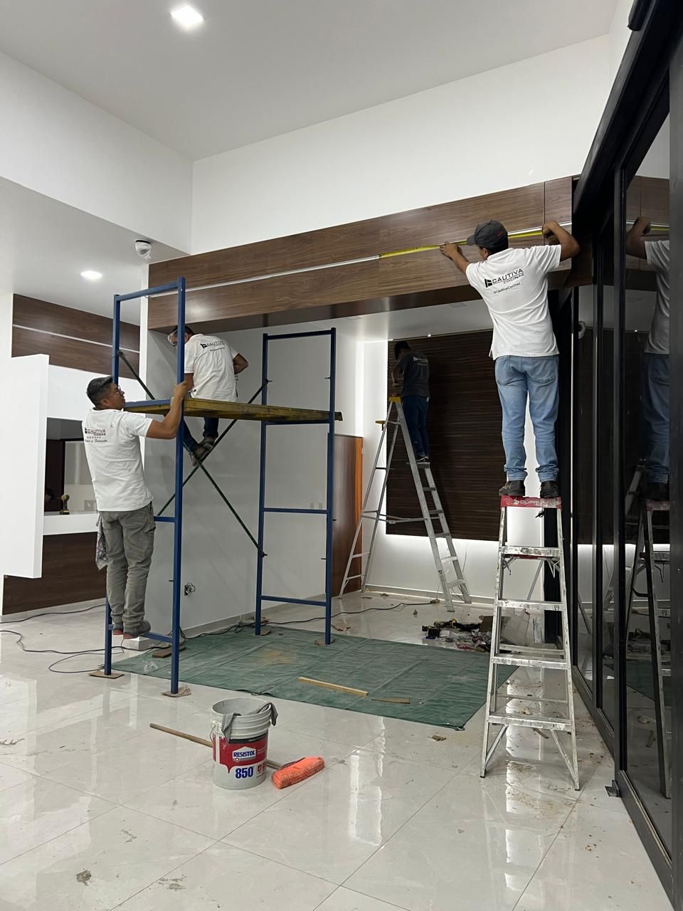 Los trabajadores instalan paneles de madera, utilizando andamios y escaleras, en un interior moderno con puertas de vidrio.