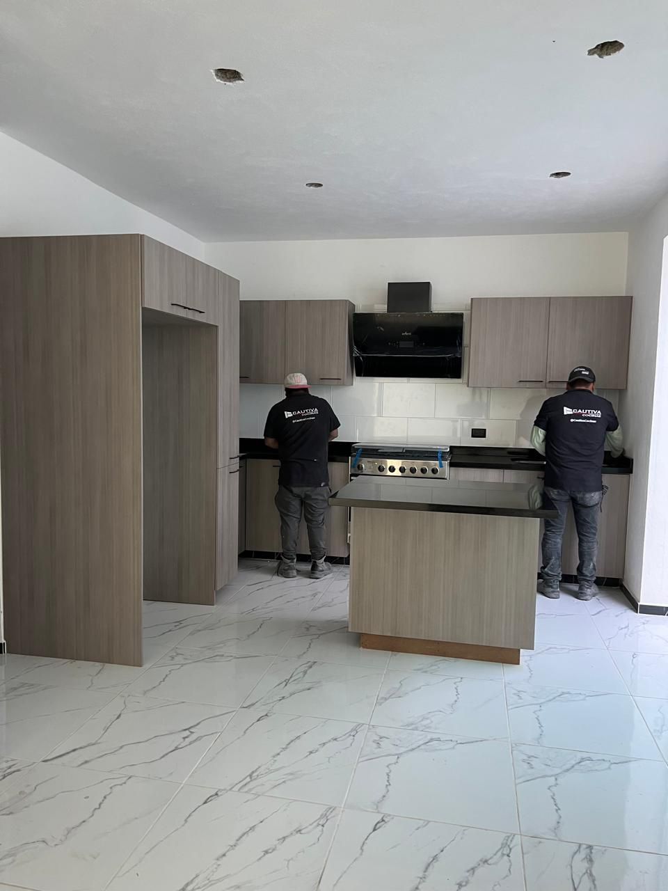 Dos trabajadores instalando nuevos gabinetes grises en una cocina con pisos de mármol blanco y electrodomésticos de acero inoxidable.