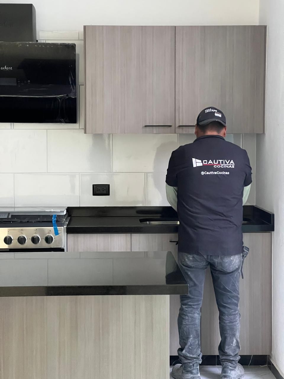 Una persona trabaja en la encimera de la cocina. La cocina tiene gabinetes de madera clara, encimeras negras y una estufa.