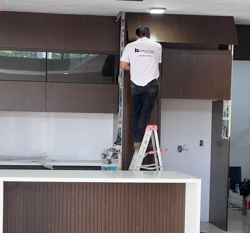 Una persona instala gabinetes en una cocina. Está en una escalera, trabajando en los gabinetes marrón oscuro.