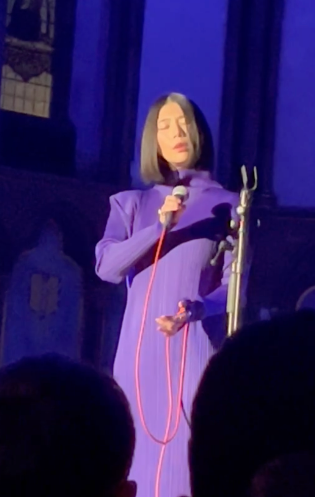 Woman in purple dress sings into a microphone on stage, short black hair, blue lighting.