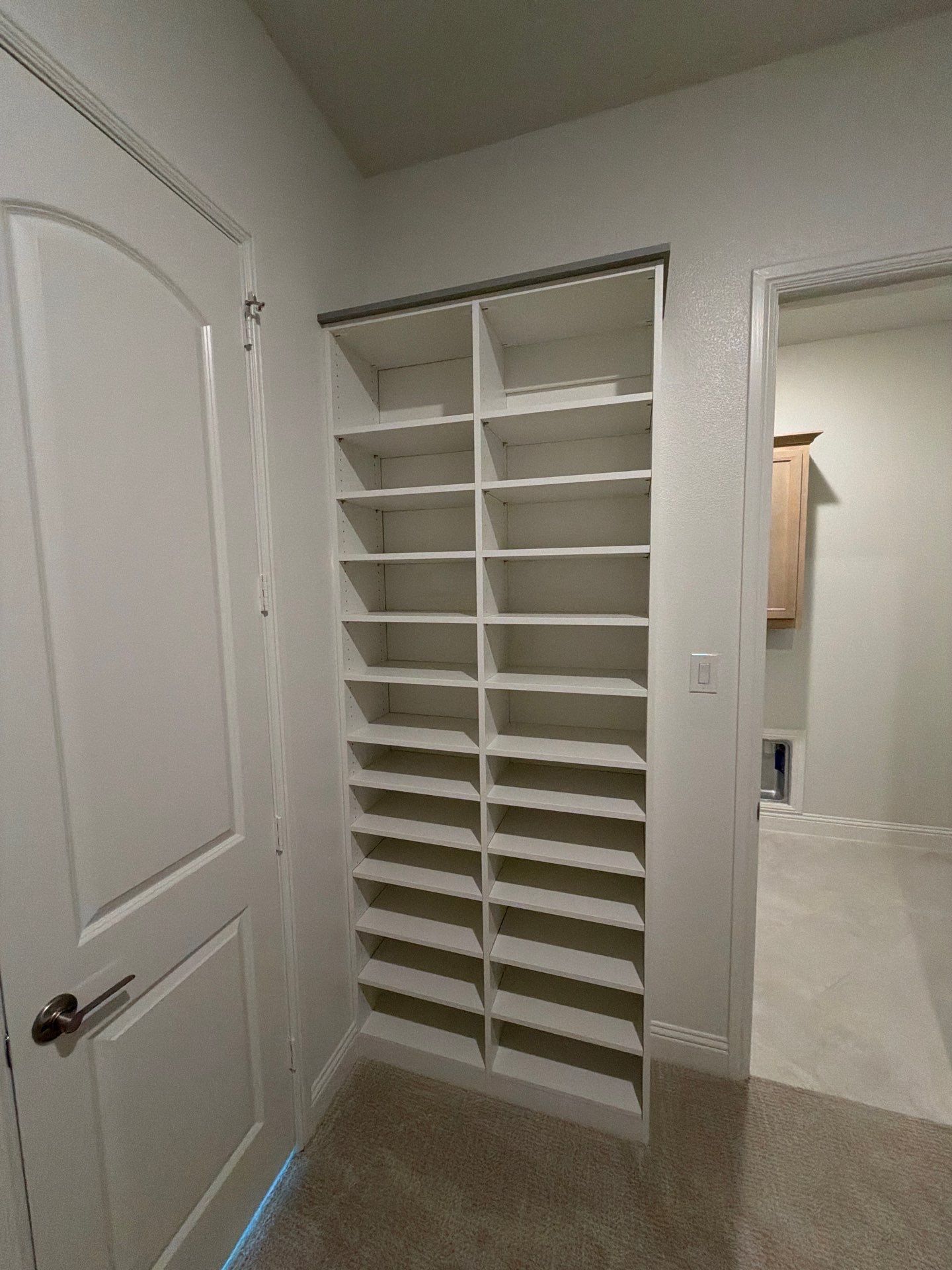White closet shelving unit in a hallway or closet, with a door on the left and an open doorway on the right.