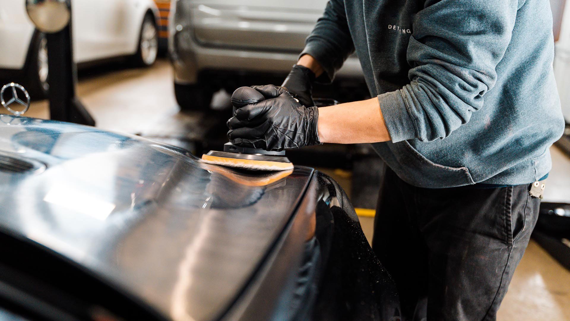 Person in gloves polishing a car's surface with an electric buffer in a garage.
