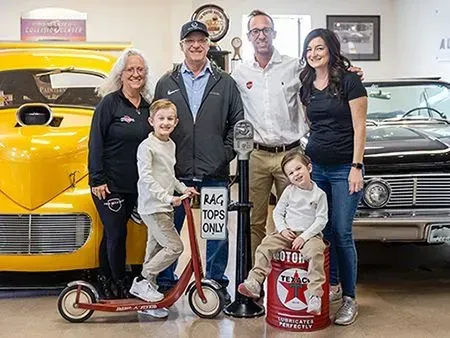 Family poses with vintage cars, a boy on a scooter, and a boy on a gas can in a garage.