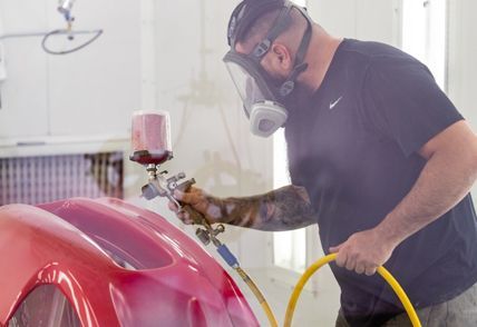 Person in respirator paints red car part in a spray booth, holding a paint gun.