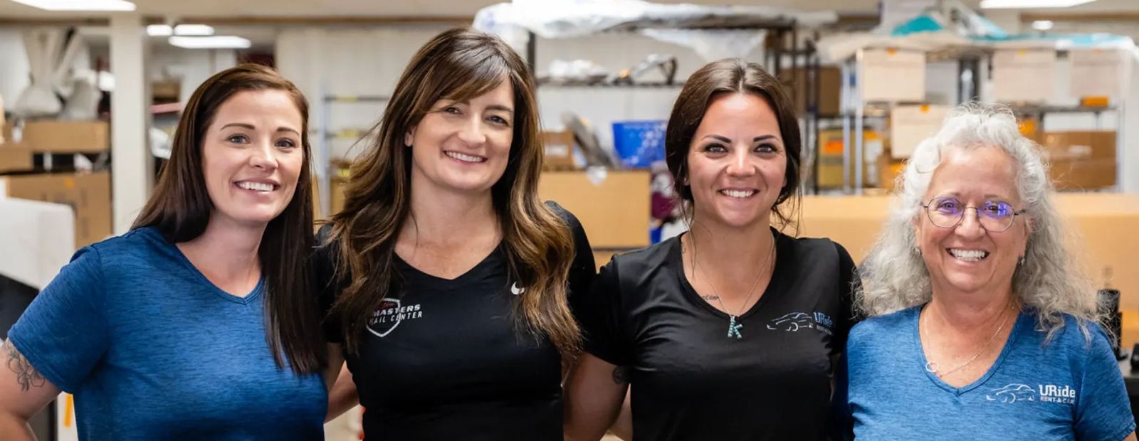 Four women smile in a workshop, wearing blue and black V-neck shirts.