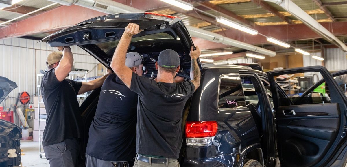 Three people standing in front of a silver SUV with the hatch open. They are smiling. Indoor setting.