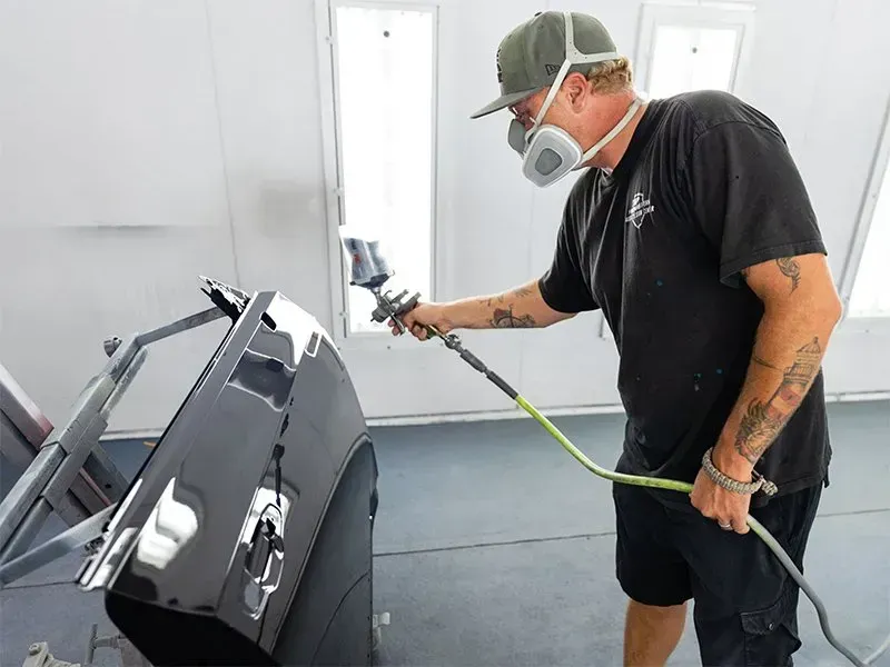 Person sprays black paint on car part, wearing respirator in a workshop.