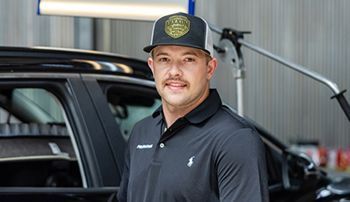 Man in a cap, smiling, stands near a black car inside a shop; a lamp is above.