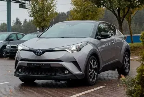 A silver Toyota C-HR parked on a paved lot with trees in the background under an overcast sky.