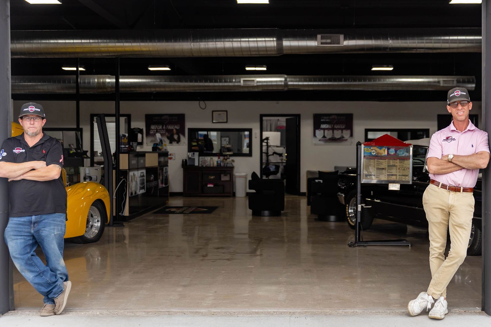 Two men standing in a car showroom entrance, with a yellow car inside and office furniture in the background.