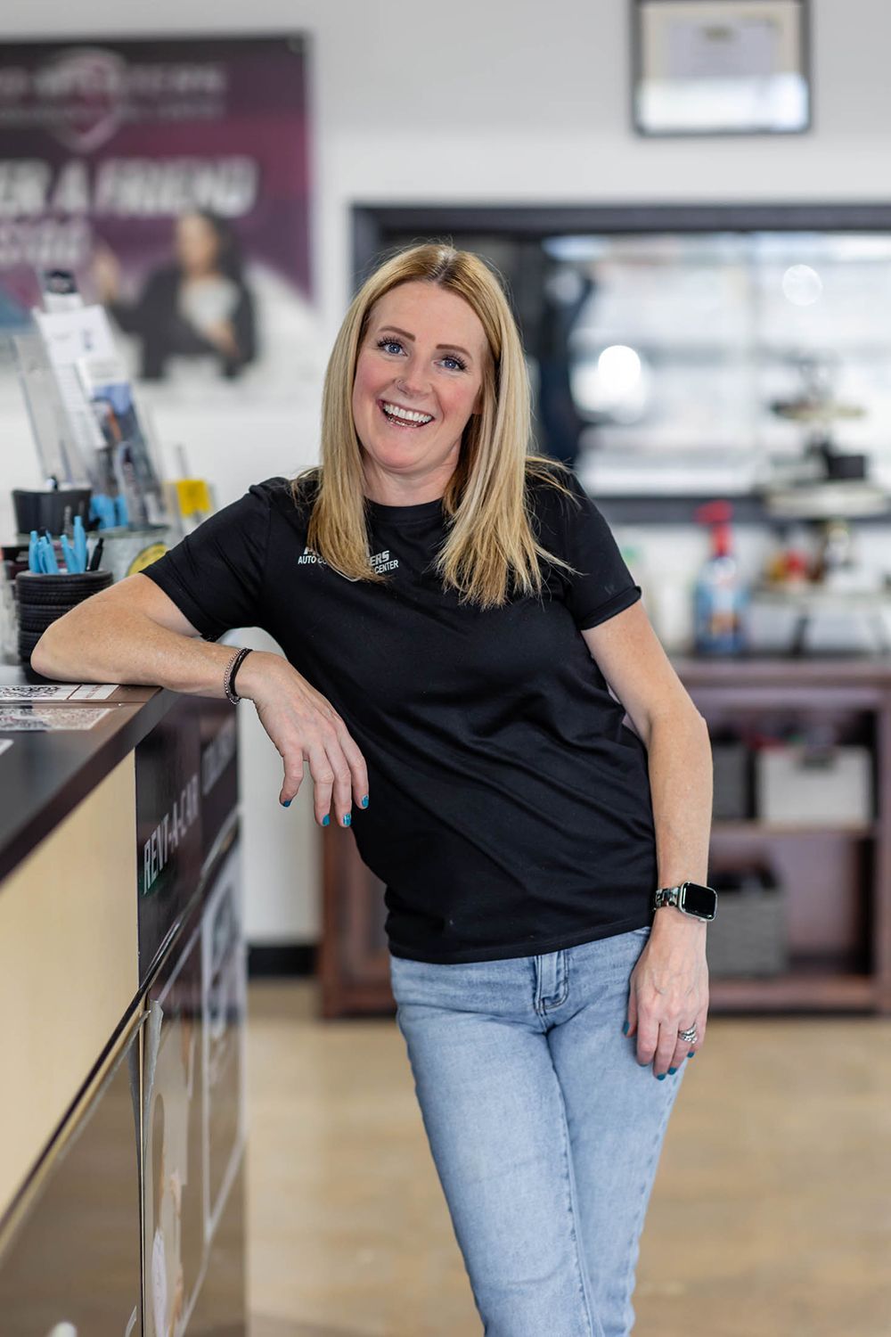Smiling woman in a black shirt and light jeans leaning on a counter in a store