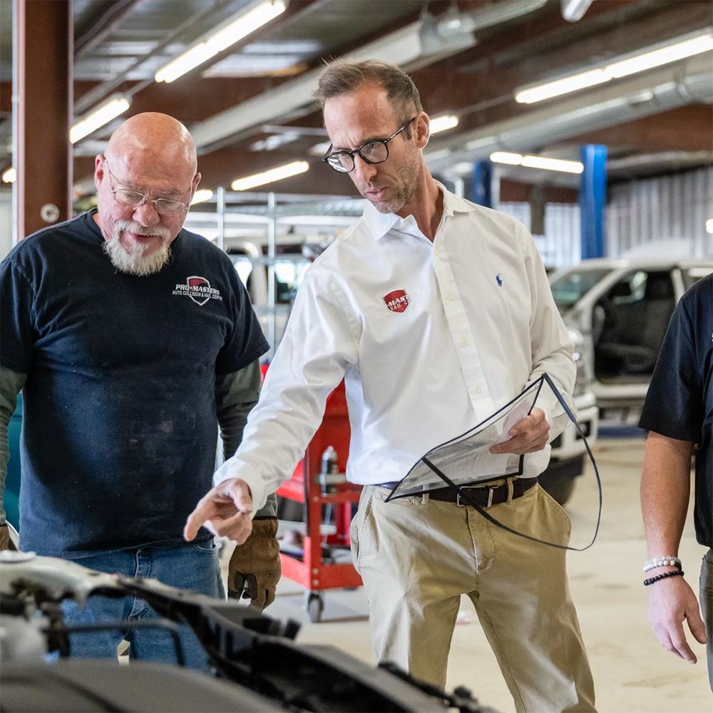 Two men inspect a car in a repair shop, one holding a clipboard and pointing at the engine.