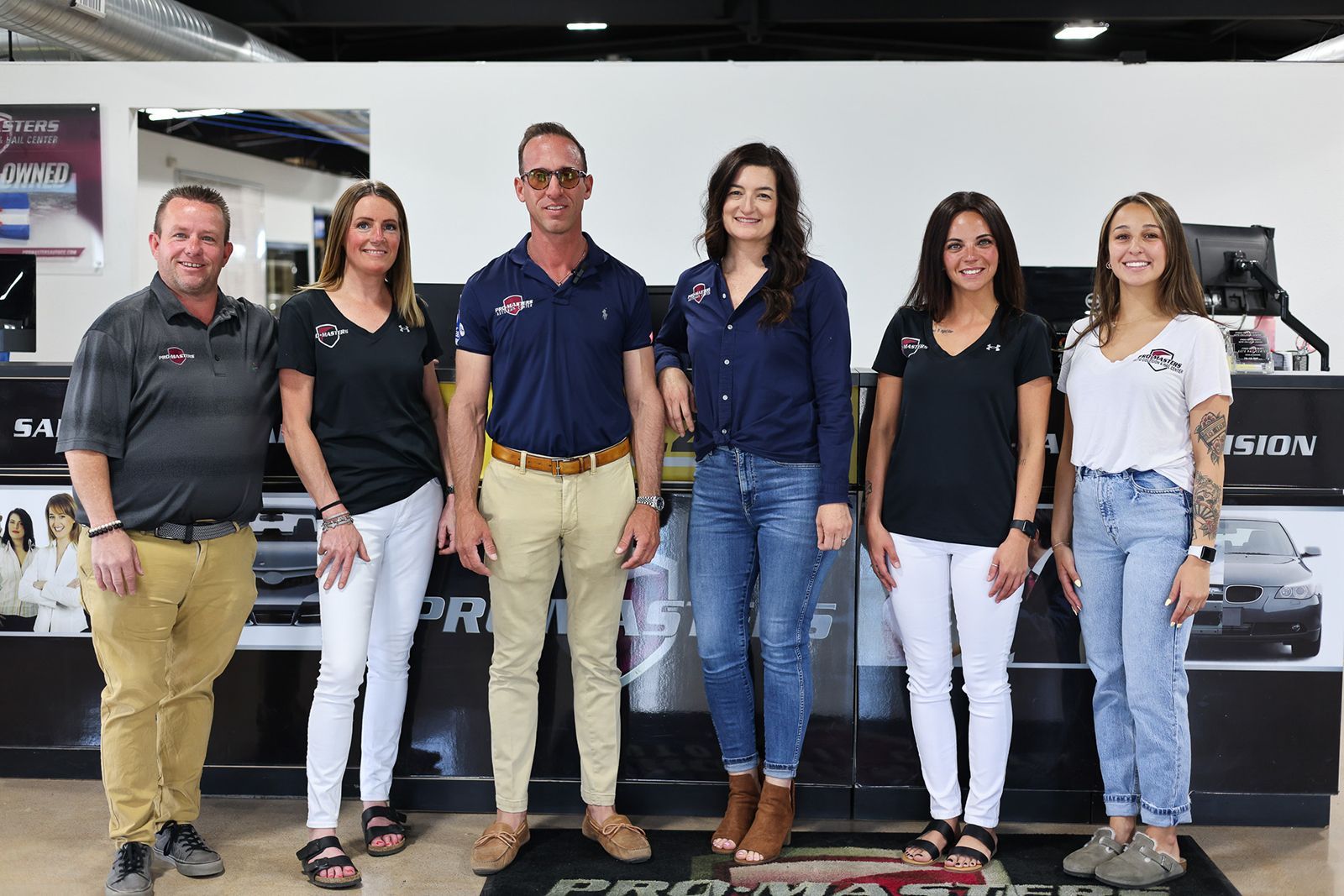 Six people posing indoors in front of a dealership display, wearing casual clothes and smiling.
