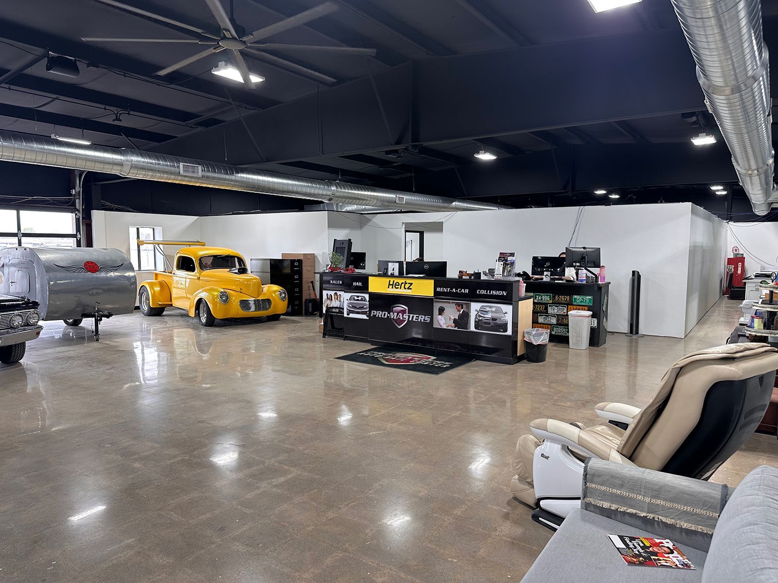 Spacious garage showroom with polished floors, classic yellow car, desks, tools, and chairs under black ceiling beams