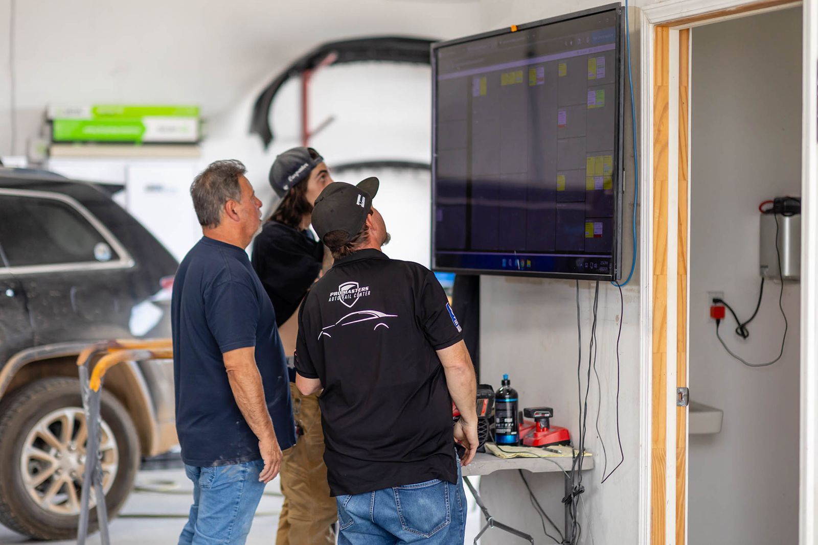 Three people viewing a wall-mounted monitor in a garage workspace