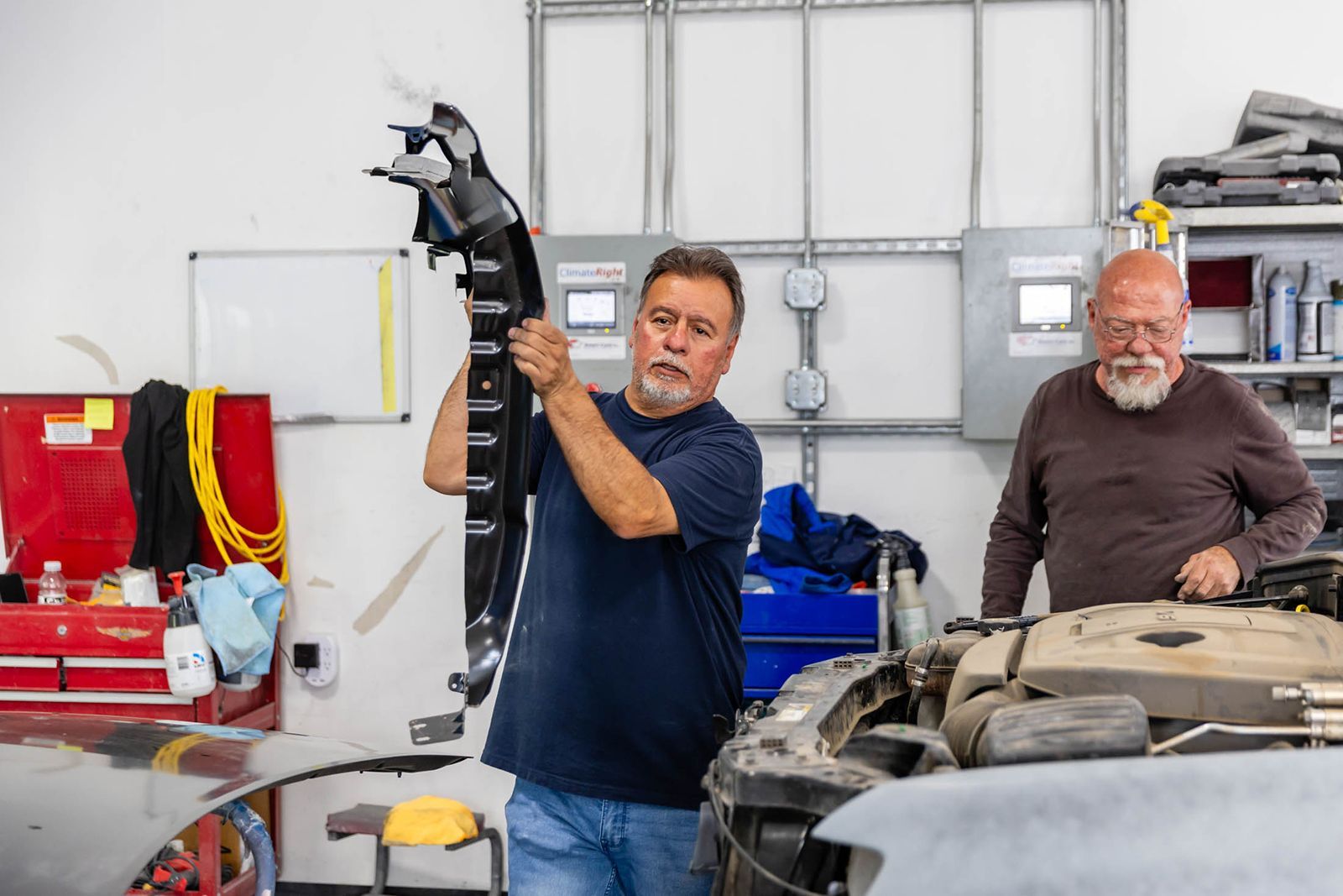 Two mechanics in a garage, one holding a car part beside an open engine bay