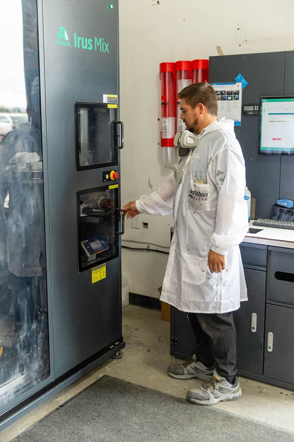 Person in a lab coat operates a lab machine beside a computer monitor in a lab.