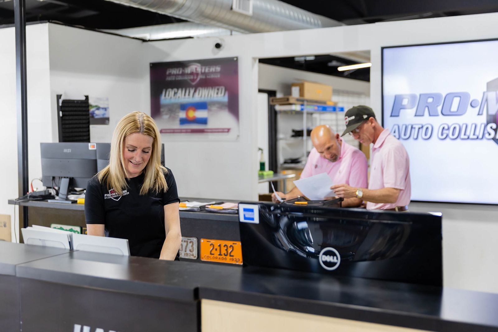 Reception desk at an auto collision shop with staff assisting customers and a black car hood in the foreground