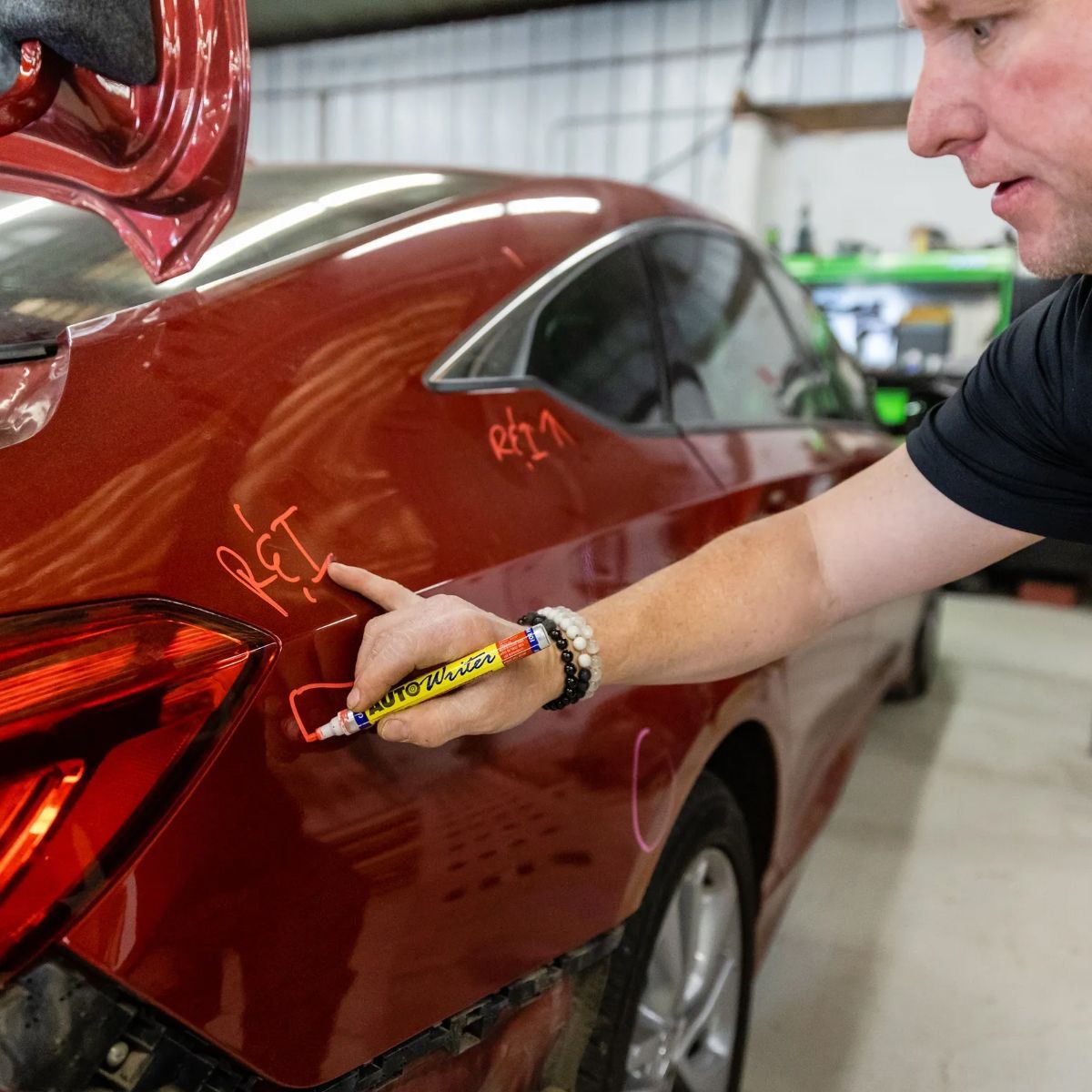 Person marking a red car’s rear quarter panel in a workshop while pointing at the damage.