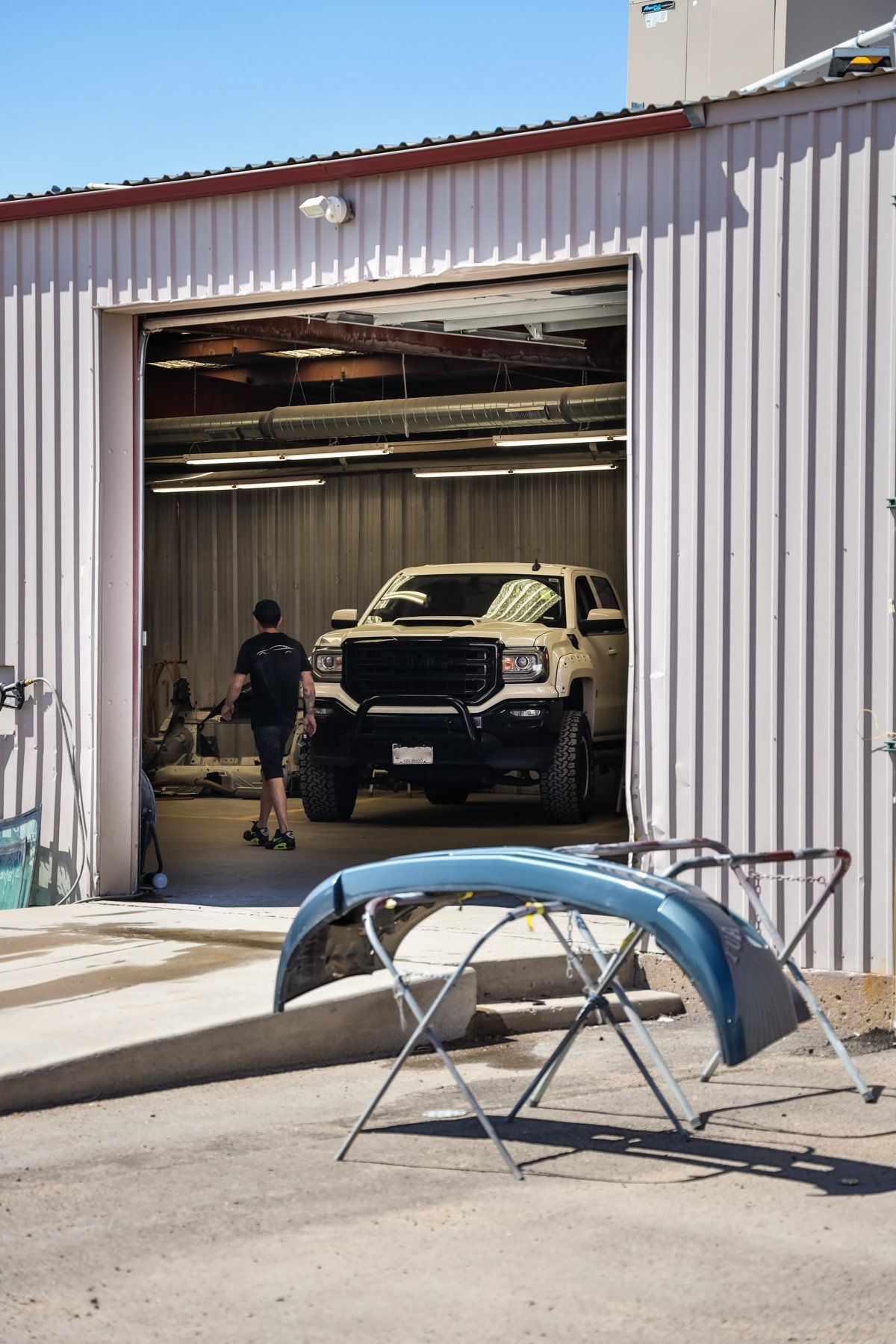A large, white truck inside a garage; a person walks in front of it. A bumper is on a stand in front of the garage.