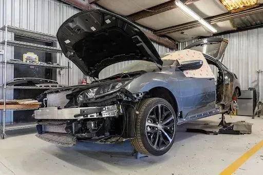 A partially disassembled gray car in a repair shop with its hood and door open.