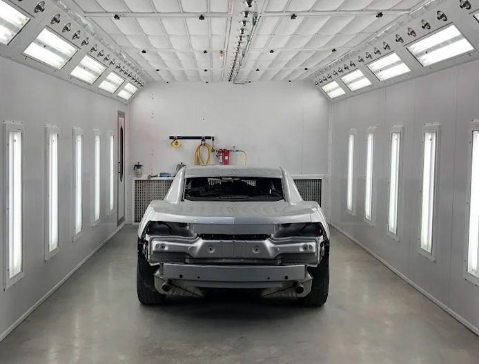 White pickup truck inside a bright auto paint booth with overhead lights and open wall panels