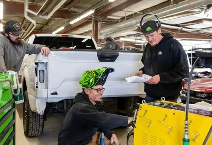 Three people working on a white pickup truck in a garage, with tools and equipment nearby.