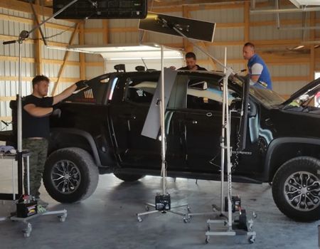 Black pickup truck in a workshop, lifted by jacks while three people work on it.