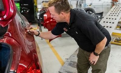 Man in black shirt examines dent on red car's fender with a pencil in a repair shop.