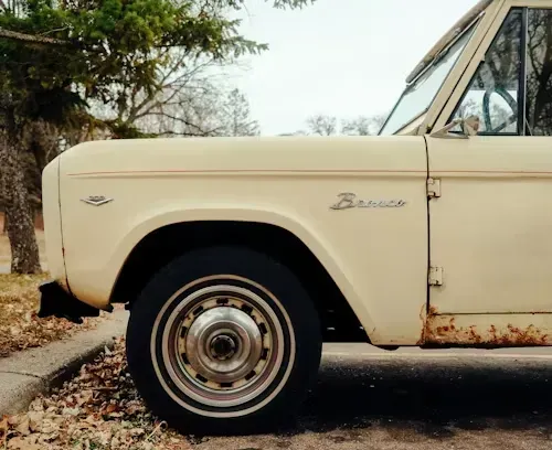 Off-white vintage Ford Bronco parked on the side of a road, with visible rust and a tree in the background.