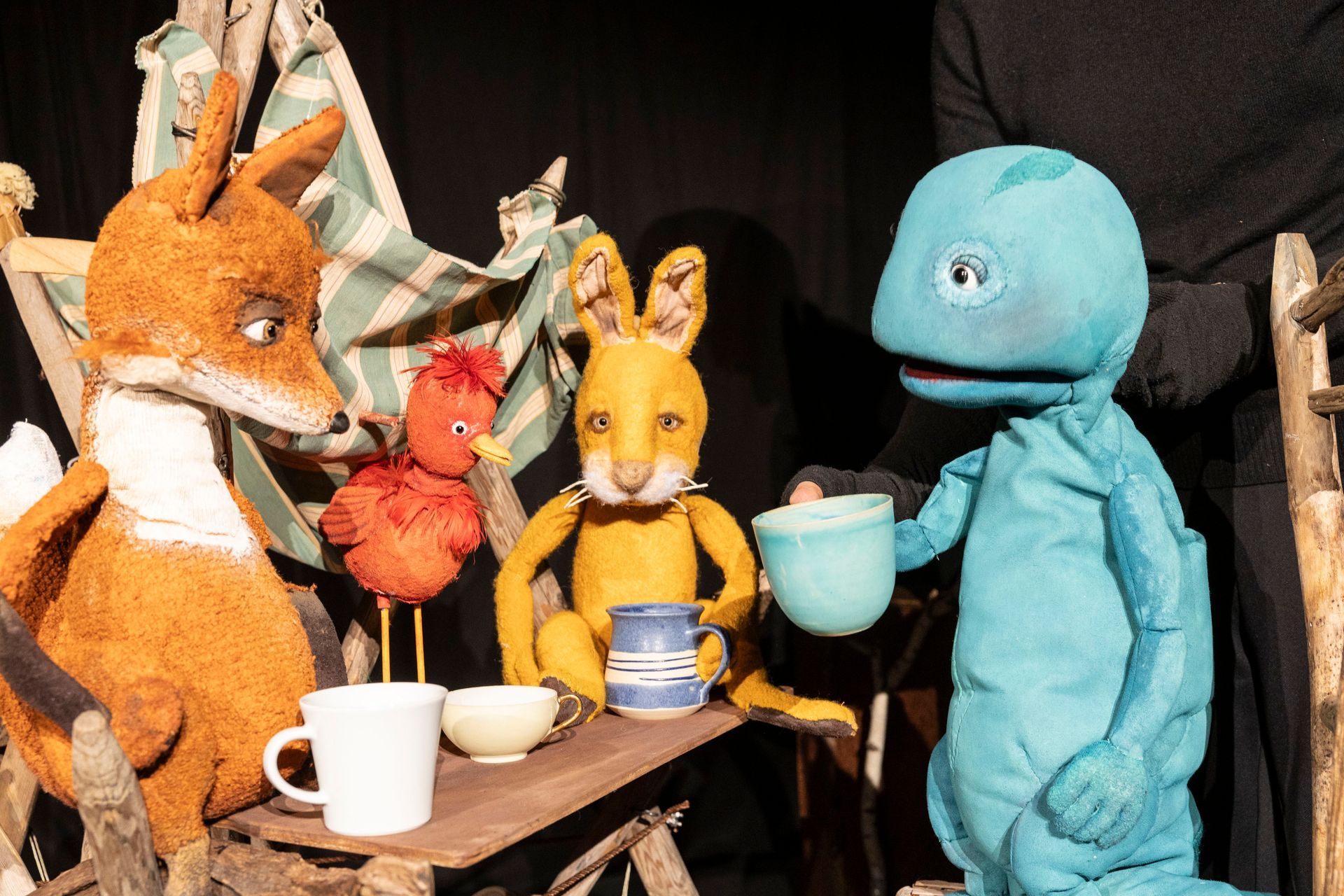 Puppenspiel: Fuchs-, Vogel-, Kaninchen- und Eidechsenpuppen an einem Tisch. Die Figuren scheinen sich beim Teetrinken zu unterhalten.